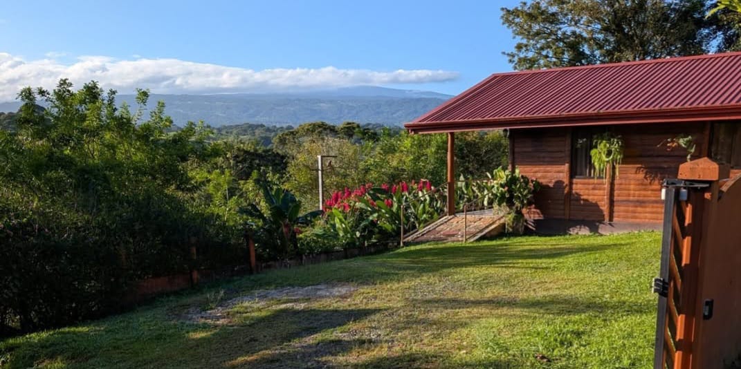 Wooden cabin with red roof overlooking forested hills and a distant volcano